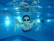 Ein Mädchen mit Schwimmbrille taucht unter Wasser freudig auf die Kamera zu.