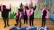 Fünf Kinder tanzen in schwarz gekleidet mit rosa Federboa und rosa Flamingokopf einen fröhlichen Tanz. Sie reißen die Arme nach oben und lachen in einer Kulisse im Meer.