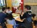 4 Schüler, eine Schülerin und eine Lehrerin sitzen vor einer geöffneten Klassentür auf dem Flur um einen Tisch und lachen herzlich. Vor ihnen auf dem Tisch liegen Spielanleitungen und in der Mitte ein Würfel.