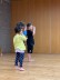 Drei Kindergartenkinder stehen in einem großen Raum. Alle tragen sportliche Kleidung. Sie führen mit ihrer rechten Hand eine Bewegung vor ihren Gesichtern aus. 