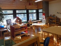 Blick in einen Gruppenraum: Im Hintergrund ist eine Fensterfront, davor sind unterschiedliche Holzmöbel und Spielmaterial zu sehen, unter anderem ein Puppenhaus und viele Bücher. Die Sonne scheint in den Raum herein.