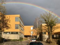 Ansicht des Haupteingangs der Sekundarstufe I. Das Gebäude wird von der tiefstehenden Sonne angestrahlt, darüber spannt sich vor grauem Himmel ein kräftiger Regenbogen.
